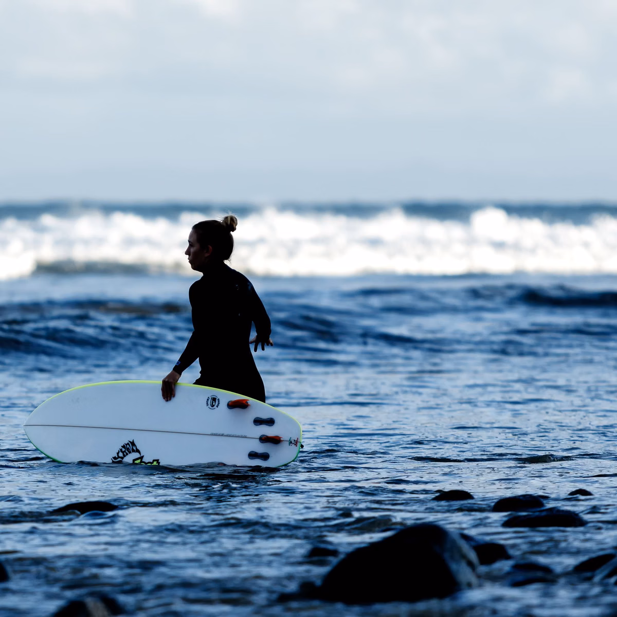 A surfer in a wetsuit wading out over cobbles with a surfboard in Malibu