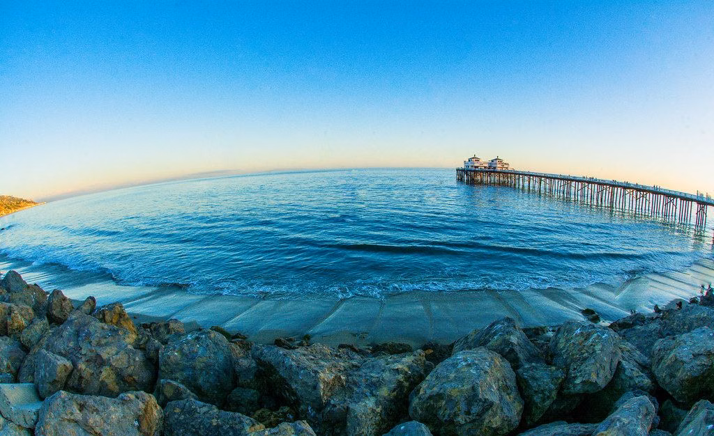 The Malibu Pier stretching out over calm water at sunset, seen from the rocks on shore