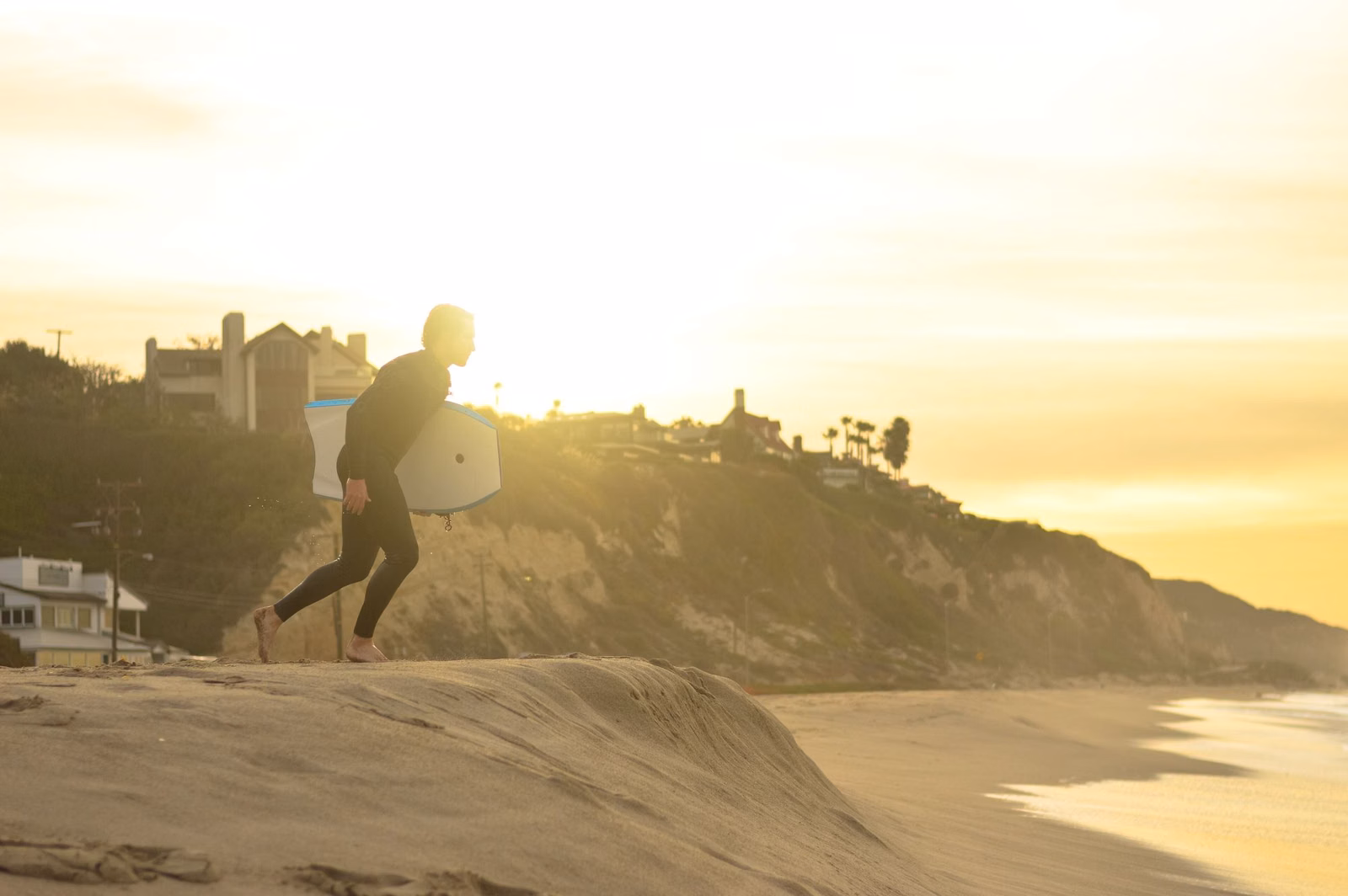 A surfer with a board walking along a Malibu beach in late-day sun