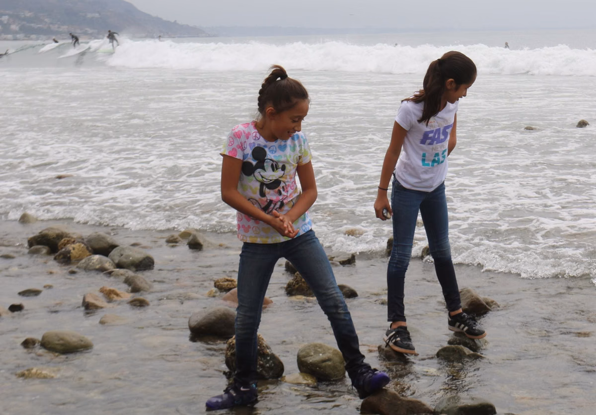 Two girls exploring the cobble tidepools at Surfrider Beach while surfers ride waves behind them